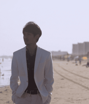 Man in a tanned suit at the beach. His jacket is open, with a dark, patterned shirt beneath. He looks back towards the tall buildings encircling the beach.